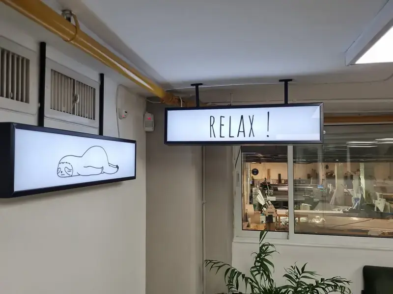 Two ceiling-hung LED lightboxes in an office corridor, one reading RELAX and one with a sloth line drawing
